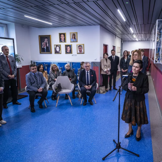 Kobieta mówi do mikrofonu, slucha ją grono osób zebranych na uroczystośći zorganizowanej na szkolnym korytarzu