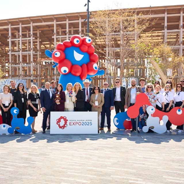 grupa osób stojących przed drewnianą konstrukcją na świeżym powietrzu. Z przodu duży baner z napisem 'EXPO 2025' oraz logo wydarzenia. Wokół nich ustawione są duże, kolorowe figury balonowe oraz kartonowe postacie w kolorach czerwonym i niebieskim, przypominające bajkowe stwory z dużymi oczami. 