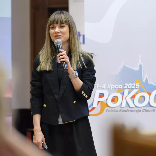 Kobieta z długimi włosami, w czarnej garsonce w czasie wsytapnie. Z tyłu wyświetla się napis konferencji PoKoChAnapisa