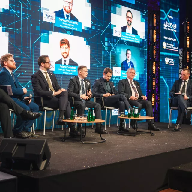 Panel ekspercki z udziałem siedmiu mężczyzn. Z tyłu na ekranie wyśietlaja się zdjęcia panelistów.