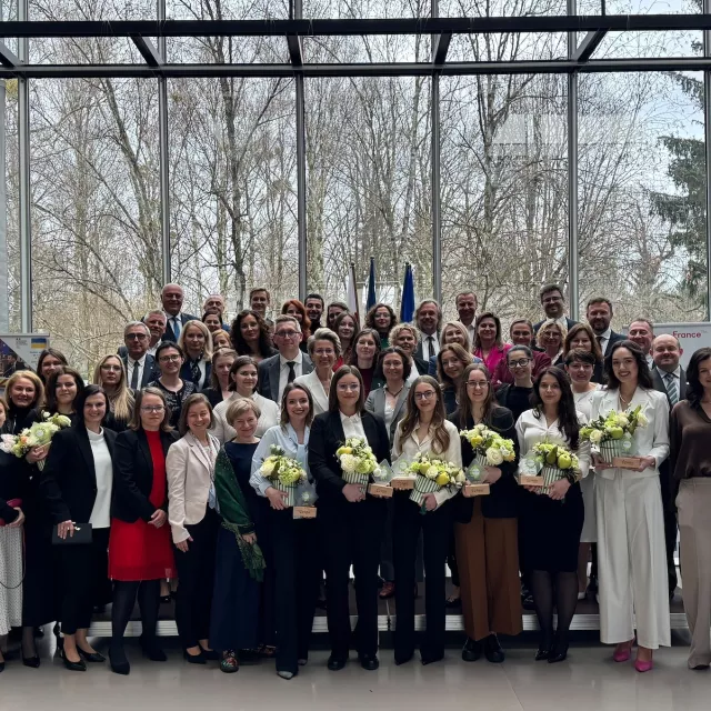 Grupa ludzi w eleganckich strojach stoi razem w pomieszczeniu z dużymi oknami w tle. Kilka kobiet osób trzyma bukiety kwiatów.